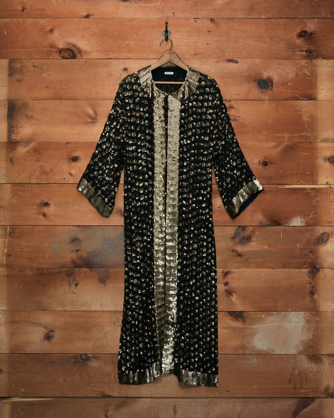 Pyrite Sequin Kaftan—a black and gold patterned long coat with three-quarter sleeves, reminiscent of a luxurious silk kaftan, hangs on a hook against a wooden wall—proudly made in India.
