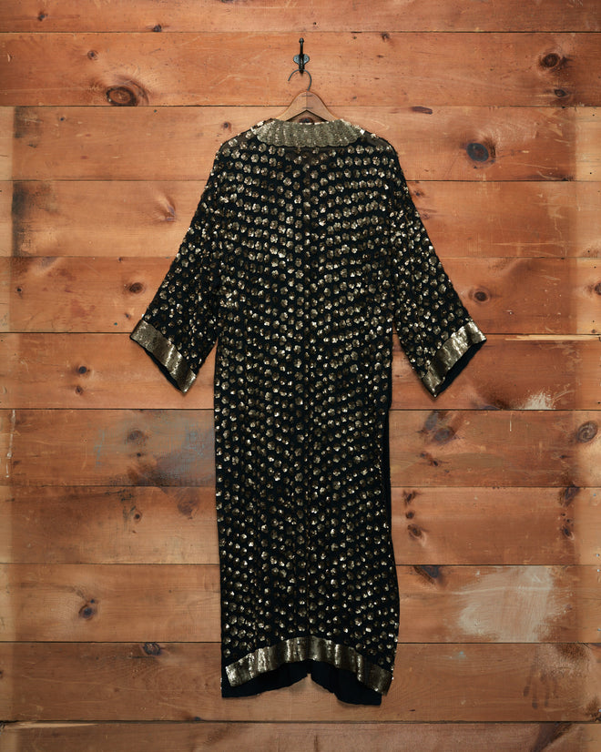 The Pyrite Sequin Kaftan in black and gold, featuring three-quarter sleeves, hangs on a wooden wall.