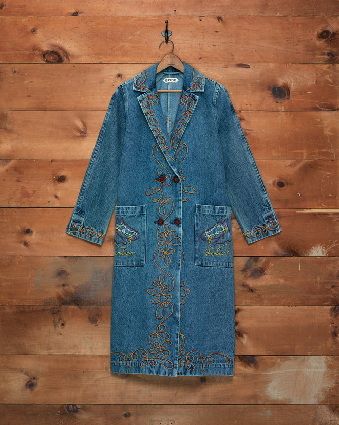 The Rawlins Coat is a long denim coat featuring colorful embroidery and distinctive horse designs on the pockets, making it a unique piece of cowboy wear that hangs beautifully on a wooden wall.