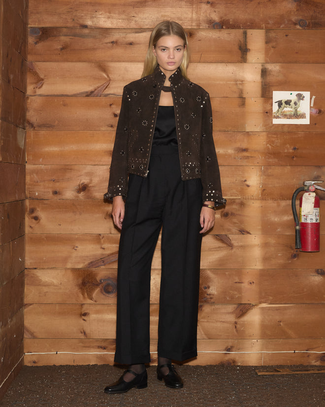 Model wearing the Studded Howard Jacket, a brown suede piece with studded details, layered over black wide-leg trousers and a cami, posed against a wood wall.