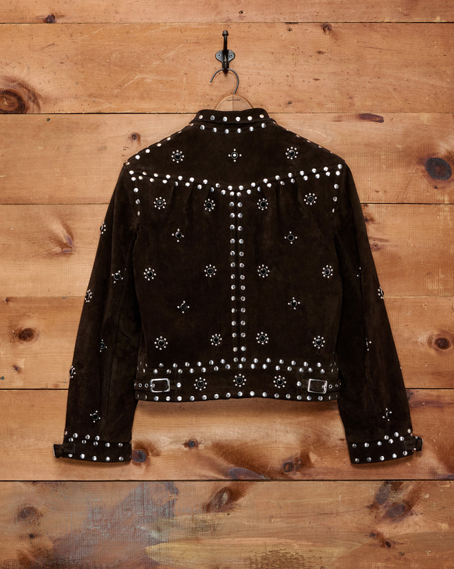 Introducing the Studded Howard Jacket—a brown suede women’s leather jacket featuring silver studs arranged in floral patterns. Hanging from a black hook on a wooden wall, this striking piece blends timeless western flair with modern elegance.