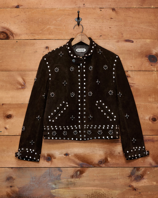 The Studded Howard Jacket, a dark brown suede piece featuring silver studs and floral patterns reminiscent of a classic women’s leather jacket, hangs on a wooden wall.