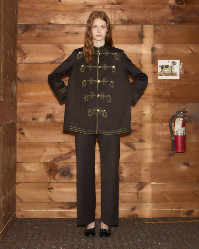 A model wears a dark, military-style Cavalry Coat in wool with gold soutache trim and matching trousers.