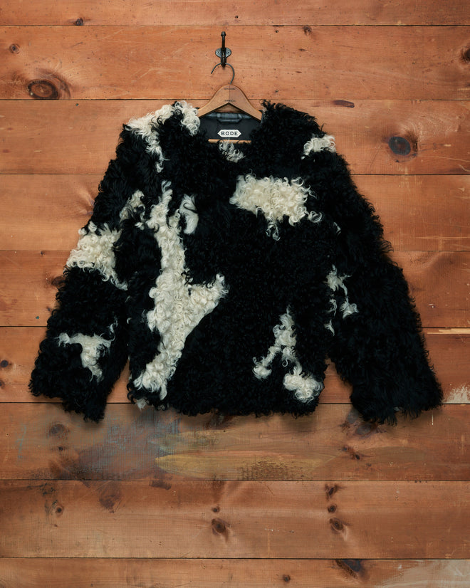 Shearling Cow Print Jacket in black and white curly faux fur, hanging on a hook against a wooden wall background.