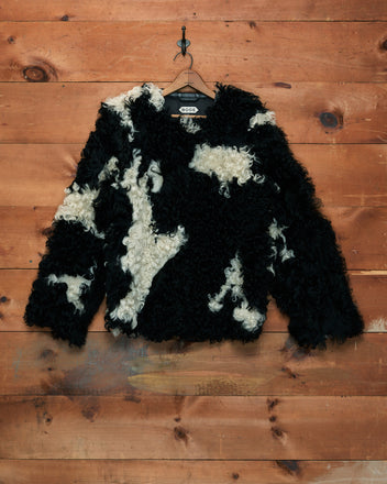 Shearling Cow Print Jacket in black and white curly faux fur, hanging on a hook against a wooden wall background.