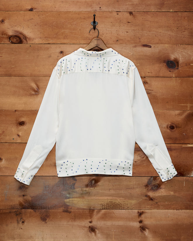The Gem Dust Long Sleeve Shirt, a white silk top with pearl studs inspired by classic Western style, is hung on a wooden wall and displayed from the back.