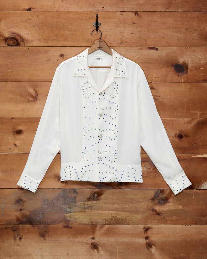 The “Gem Dust Long Sleeve Shirt,” a white silk top adorned with hand-applied rhinestone embellishments inspired by classic Western costumes, hangs on a hook against a wooden wall.
