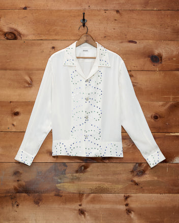 The “Gem Dust Long Sleeve Shirt,” a white silk top adorned with hand-applied rhinestone embellishments inspired by classic Western costumes, hangs on a hook against a wooden wall.