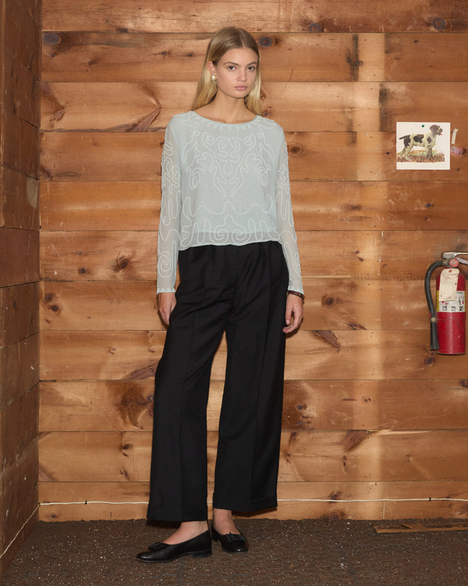 A woman in the sheer, hand-beaded Beaded Cirrus Blouse and black pants stands by a wooden wall with a fire extinguisher and photo.