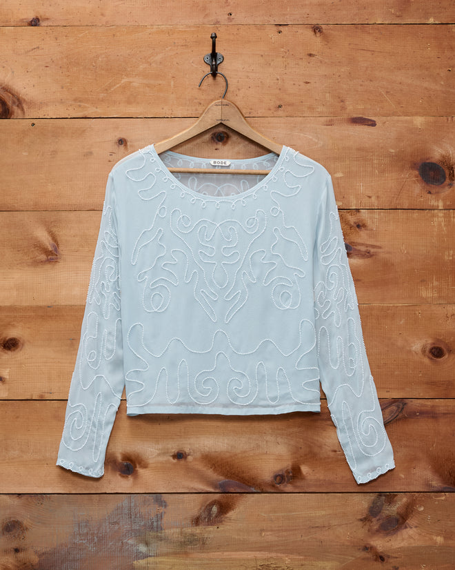 The Beaded Cirrus Blouse, a light blue long-sleeve silk top with delicate white embroidery, hangs on a wooden wall.