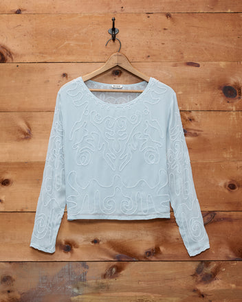 The Beaded Cirrus Blouse, a light blue long-sleeve silk top with delicate white embroidery, hangs on a wooden wall.