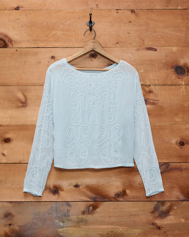 Beaded Cirrus Blouse—this white, long-sleeve overblouse features intricate beaded and embroidered patterns, displayed on a wooden hanger against a wooden wall.