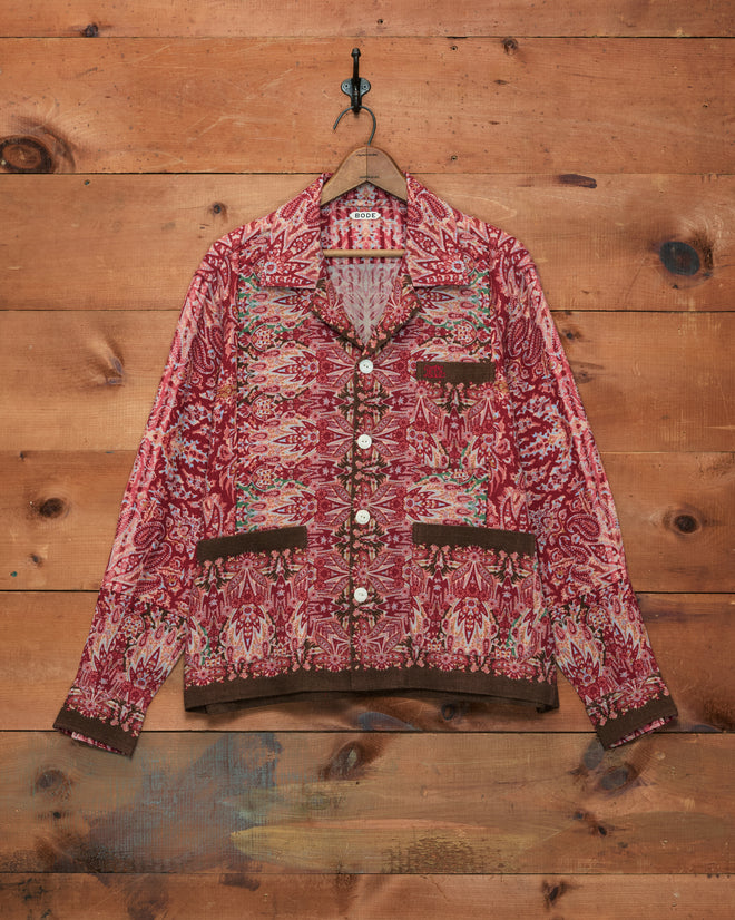 Arrowleaf Long Sleeve Shirt in a red paisley pattern with brown trim, inspired by traditional Kashmiri shawls, displayed hanging on a wooden wall.