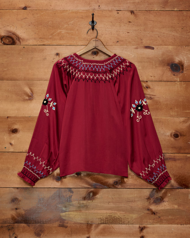A vibrant red, long-sleeve silk Evangeline Top with colorful embroidered patterns on the chest and sleeves—reminiscent of a classic 1970s blouse—hanging elegantly on a hook.
