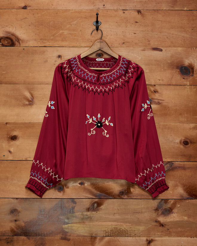 The Evangeline Top is a maroon, long-sleeve 1970s blouse featuring colorful embroidery, displayed hanging on a wooden wall.