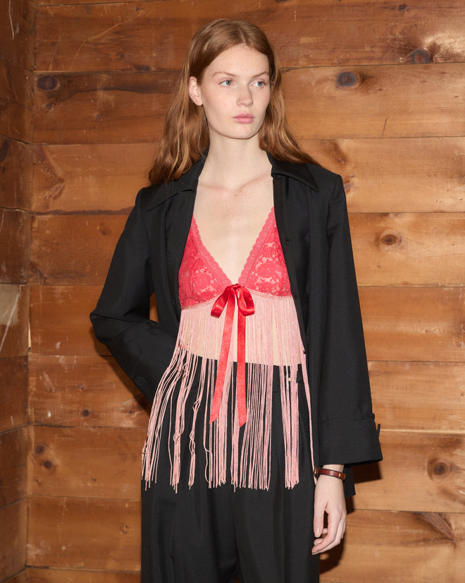 Woman in a black suit with the Jules Top, a red lace fringed bralette, standing against a wooden wall and looking to the side.