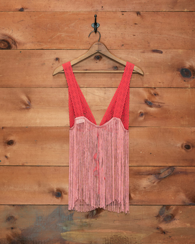 Jules Top—a red fringed sleeveless top displayed on a hanger against a wooden wall background, reminiscent of a chic fringed bralette.