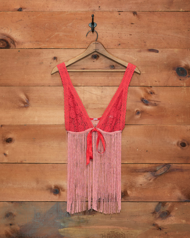The Jules Top, a red lace tank with long fringe reminiscent of a fringed bralette, hangs on a wooden hanger against a rustic wooden wall.