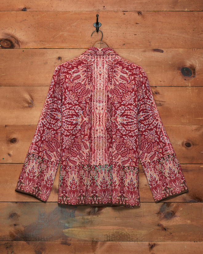 Arrowleaf Blouse, featuring red and pink patterns inspired by the intricate designs often seen in Kashmiri shawls, displayed on a hanger against a wooden wall background.