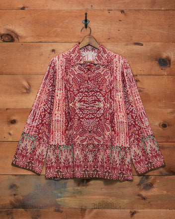 A long-sleeve collared blouse with an intricate red, pink, and beige paisley and ikat pattern, hanging on a wooden hanger against a rustic wood plank wall.