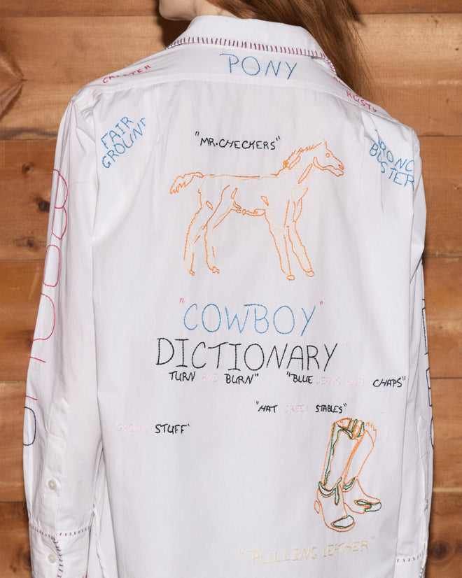 A person stands by a wood wall wearing the Rodeo Dictionary Long Sleeve Shirt, hand-embroidered from 100% cotton poplin and adorned with playful doodles, words, and a horse drawing on the back.