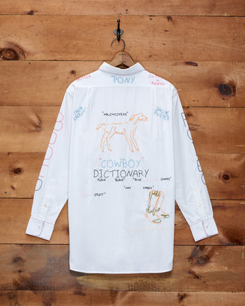 The Rodeo Dictionary Long Sleeve Shirt, made from 100% cotton poplin, hangs on a hook and showcases hand-embroidered Western doodles and words—including horses and boots—for a playful cowboy-inspired accent in any wardrobe.