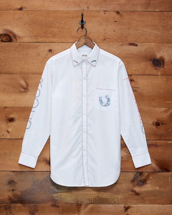 Rodeo Dictionary Long Sleeve Shirt featuring a white collar, hand-embroidered red stitching, a horseshoe motif on the pocket, and text detailing on both sleeves; crafted from 100% cotton poplin and displayed on a hanger.
