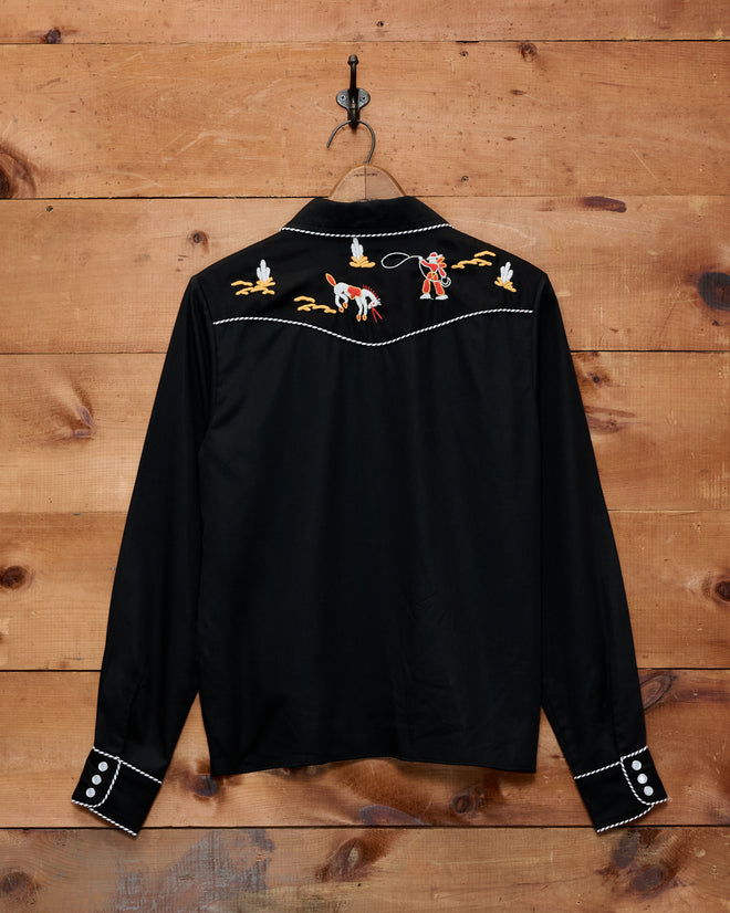 Checkers Western Blouse—a 1950s-inspired black cowboy shirt featuring embroidered cowboy, horse, and cacti on the back; displayed hanging on a wood wall.
