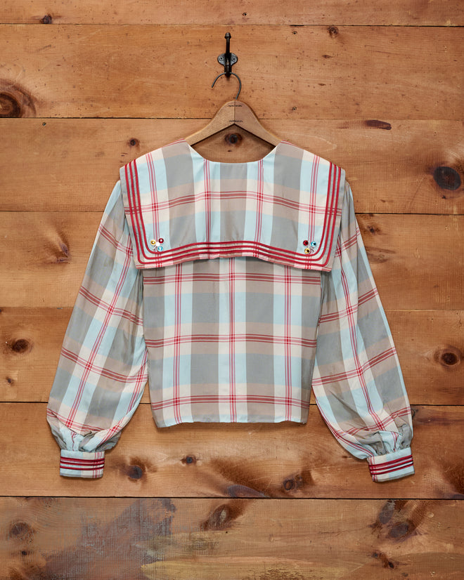 The Canyon Plaid Blouse, featuring a 1920s-inspired plaid design with long sleeves and a sailor collar, hangs on a hook against a wooden wall.