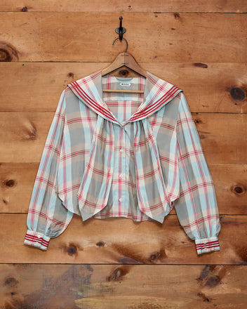 The Canyon Plaid Blouse, featuring a sheer plaid fabric, vintage-inspired 1920s sailor collar, and red accents, is displayed hanging on a wooden wall.
