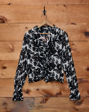 The Appaloosa Blouse, crafted from black and white patterned silk crepe with ruffled details inspired by Appaloosa spots, hangs on a wooden wall with a metal hook.