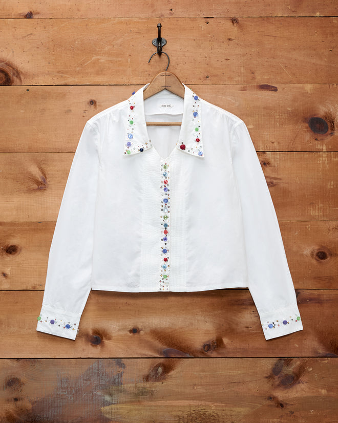 The Button Jar Blouse, a white long-sleeve shirt crafted from 100% cotton, features floral embroidery adorning the collar, front, and cuffs. Displayed on a wooden wall, this timeless blouse offers an adjustable fit and delicate details for classic elegance.