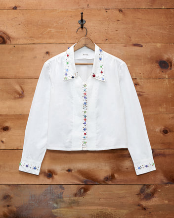 The Button Jar Blouse, a white long-sleeve shirt crafted from 100% cotton, features floral embroidery adorning the collar, front, and cuffs. Displayed on a wooden wall, this timeless blouse offers an adjustable fit and delicate details for classic elegance.