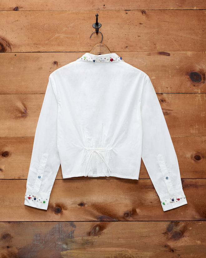 The Button Jar Blouse, a white long-sleeve top with floral embroidery and an adjustable fit blouse design, crafted from 100% cotton, is displayed from the back as it hangs on a hook against a wooden wall.