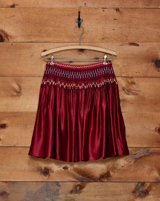 The Evangeline Skirt—a red silk piece featuring intricate blue and red embroidery with delicate chain-stitch detailing—hangs on a wooden hanger against a wood-paneled wall.