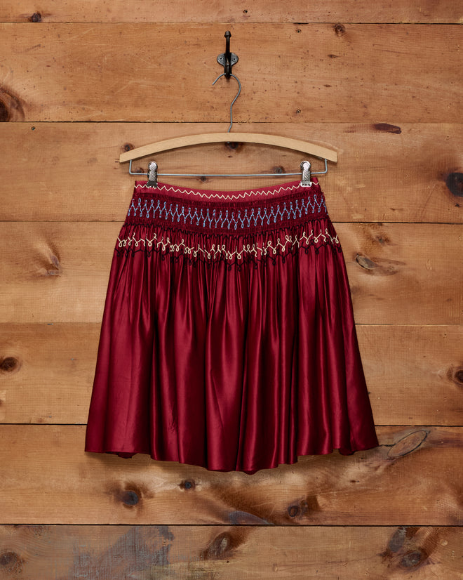 The Evangeline Skirt, crafted from red silk with a knee-length cut, features a smocked waistband and delicate chain-stitch detailing, displayed on a wooden hanger against a wooden wall.