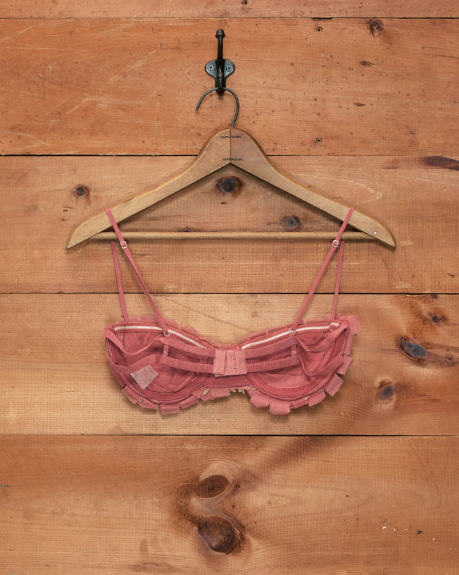The Prairie Bra, a pink lace design inspired by 1920s bralettes, hangs on a wooden hanger against a wooden plank wall.