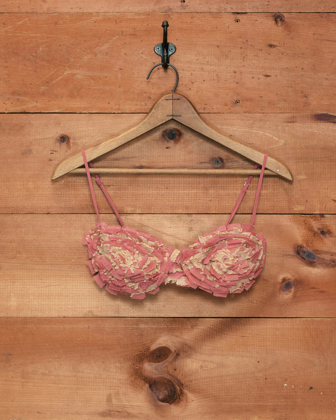 The Prairie Bra, a whimsically styled pink bra reminiscent of a 1920s bralette and crafted from deli meat, hangs on a wooden hanger against a wooden wall with a metal hook.