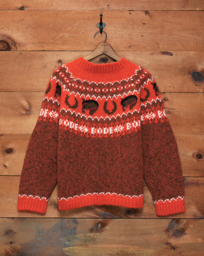 The Corral Sweater—a red and brown knit with a patterned yoke—hangs on a hook against a wooden wall. Expertly crafted in Peru from cozy alpaca, this sweater blends warmth and timeless style.