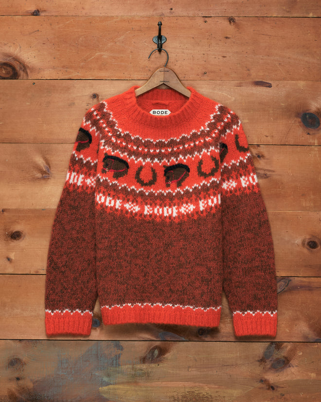 Corral Sweater—red and brown patterned alpaca knit with black abstract shapes, made in Peru, hanging on a decorative hook against a wooden wall.
