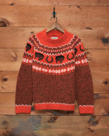 Corral Sweater—red and brown patterned alpaca knit with black abstract shapes, made in Peru, hanging on a decorative hook against a wooden wall.