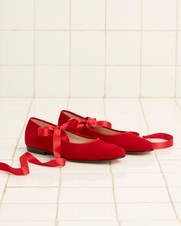 A pair of Musette Flats - Chili with satin ribbons rests on a white tiled floor.