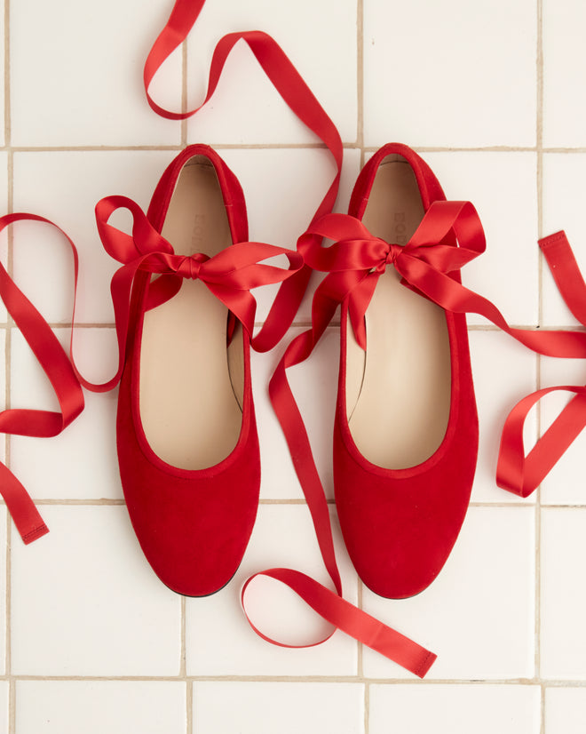 A pair of Musette Flats - Chili with satin ribbons rests on a white tiled floor.