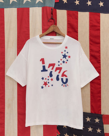 The 1776 Tee, a white graphic shirt with red and blue stars, made from vintage-weight cotton, hangs on a hanger in front of an American flag.