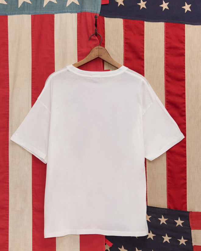 The 1776 Tee, a plain white vintage-weight cotton T-shirt, hangs on a wooden hanger in front of an American flag backdrop.