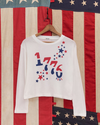 The 1776 Long Sleeve Tee, a white women’s shirt in vintage-weight cotton with a bold 1776 graphic and stars, is displayed against vintage American flag fabric.