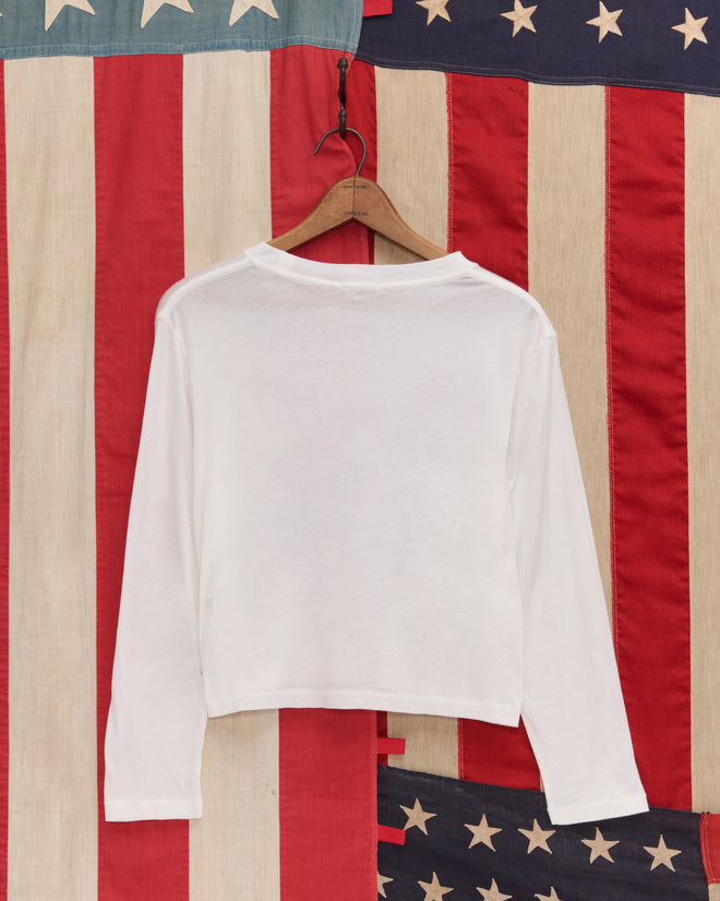 The 1776 Long Sleeve Tee, crafted from vintage-weight cotton, hangs on a hanger against an American flag backdrop.