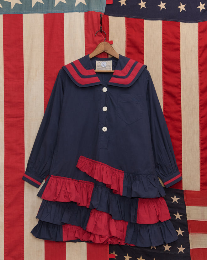 The Centennial Dress - Blue/Red, with red ruffles and a sailor collar, hangs on a wooden hanger in front of American flags, evoking classic Fourth of July styles.