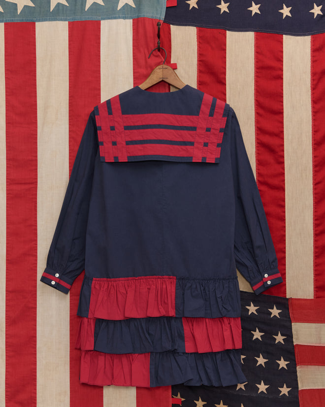 Centennial Dress - Blue/Red featuring navy fabric with red stripes and ruffled tiers, inspired by antique Liberty dress styles, displayed in front of large American flags.