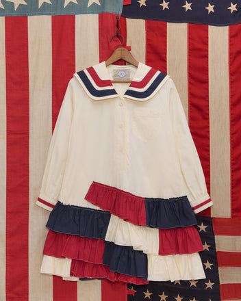 The Centennial Dress - Multi features red, white, and blue ruffles in a cream sailor style, displayed before American flag-inspired fabric—a timeless tribute to Liberty dress styles and an ideal choice for Fourth of July celebrations.